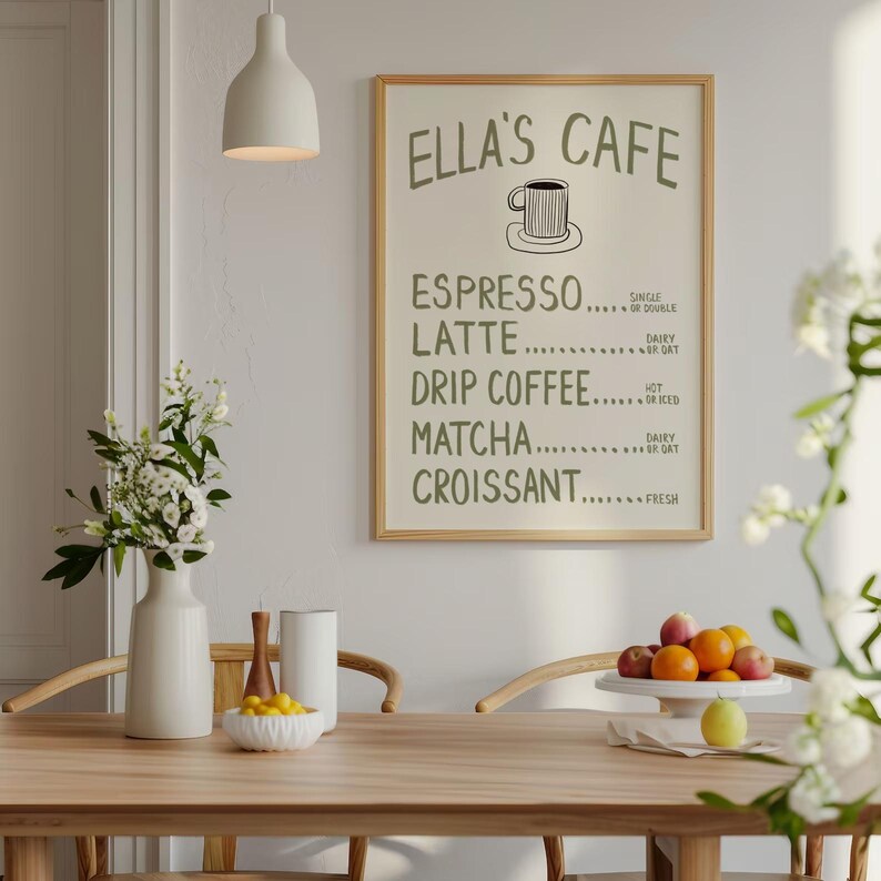 Puede incluir: Un letrero enmarcado para "Ella's Cafe" cuelga de una pared blanca, que enumera las opciones de caf&eacute; y pasteler&iacute;a. El letrero est&aacute; en un marco de madera clara. Un jarr&oacute;n blanco con flores y una mesa de madera con fruta completan la escena.