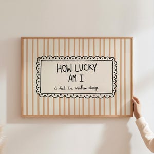 May include: A framed wall art piece with a beige and white striped background. The artwork features a black and white scalloped border around the text "HOW LUCKY AM I to feel the weather change."