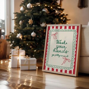 May include: A framed sign with the text "Wash your hands ya filthy animal" in green script, framed with red and white stripes and a scalloped border. The sign is propped up on a wooden floor, with a Christmas tree and wrapped gifts in the background.