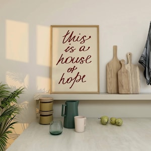 May include: A framed print with the text 'this is a house of hope' in red cursive font on a cream background. The print is hanging on a white wall above a shelf with a green pitcher, a white mug, and three green pears on a white countertop.