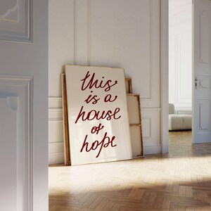 May include: A white canvas with the words 'This is a house of hope' written in red cursive font. The canvas is leaning against a white wall in a room with a wooden floor.