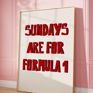 Op de afbeelding: Een ingelijste poster met de tekst "SUNDAYS ARE FOR FORMULA 1" in vette rode letters met een zwarte omlijning. De poster leunt tegen een roze muur en een houten vloer.