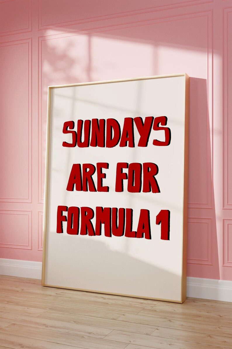 Op de afbeelding: Een ingelijste poster met de tekst "SUNDAYS ARE FOR FORMULA 1" in vette rode letters met een zwarte omlijning. De poster leunt tegen een roze muur en een houten vloer.