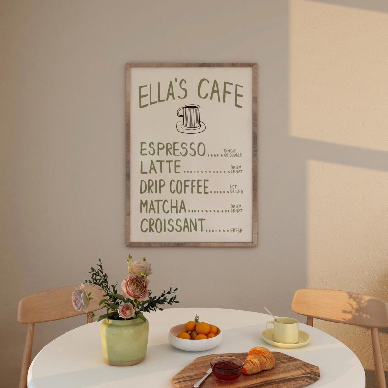 Puede incluir: Un letrero enmarcado con el texto "ELLA'S CAFE" y un men&uacute; de caf&eacute; y boller&iacute;a. Una mesa est&aacute; puesta con un jarr&oacute;n de flores, un cuenco de naranjas y un cruas&aacute;n.