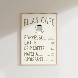 Puede incluir: Un cartel enmarcado con el texto "ELLA'S CAFE" en verde, que enumera opciones de caf&eacute; y pasteler&iacute;a. El men&uacute; incluye espresso, latte, caf&eacute; de goteo, matcha y croissant. Hay una peque&ntilde;a ilustraci&oacute;n de una taza de caf&eacute;.