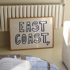 May include: A framed print with a white background and blue text that reads "EAST COAST" with a small heart symbol at the bottom right corner.