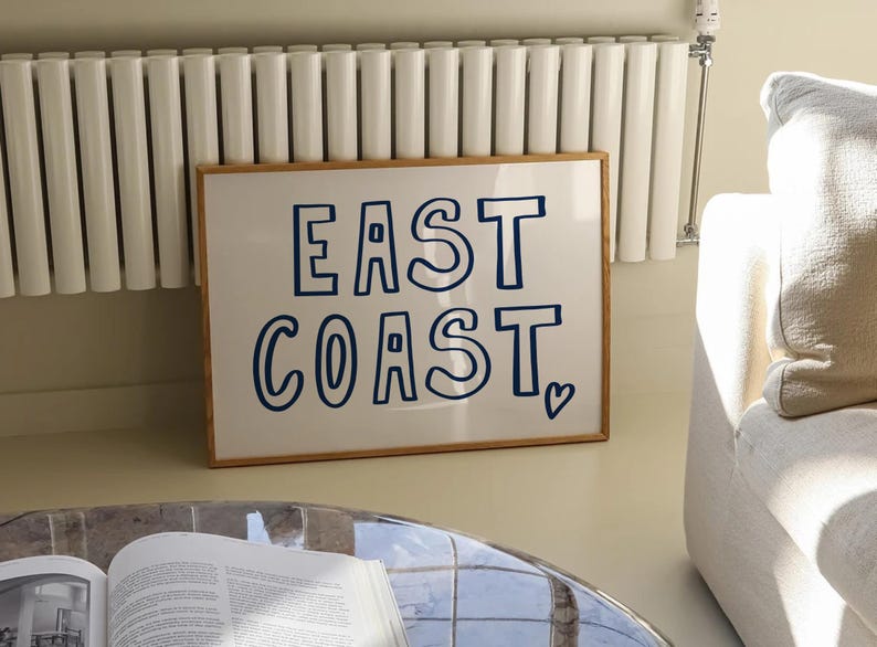 May include: A framed print with a white background and blue text that reads "EAST COAST" with a small heart symbol at the bottom right corner.