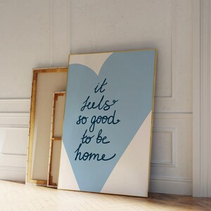 May include: A blue and white canvas print with a gold frame. The print features a blue heart shape with the text "it feels so good to be home" written in black.