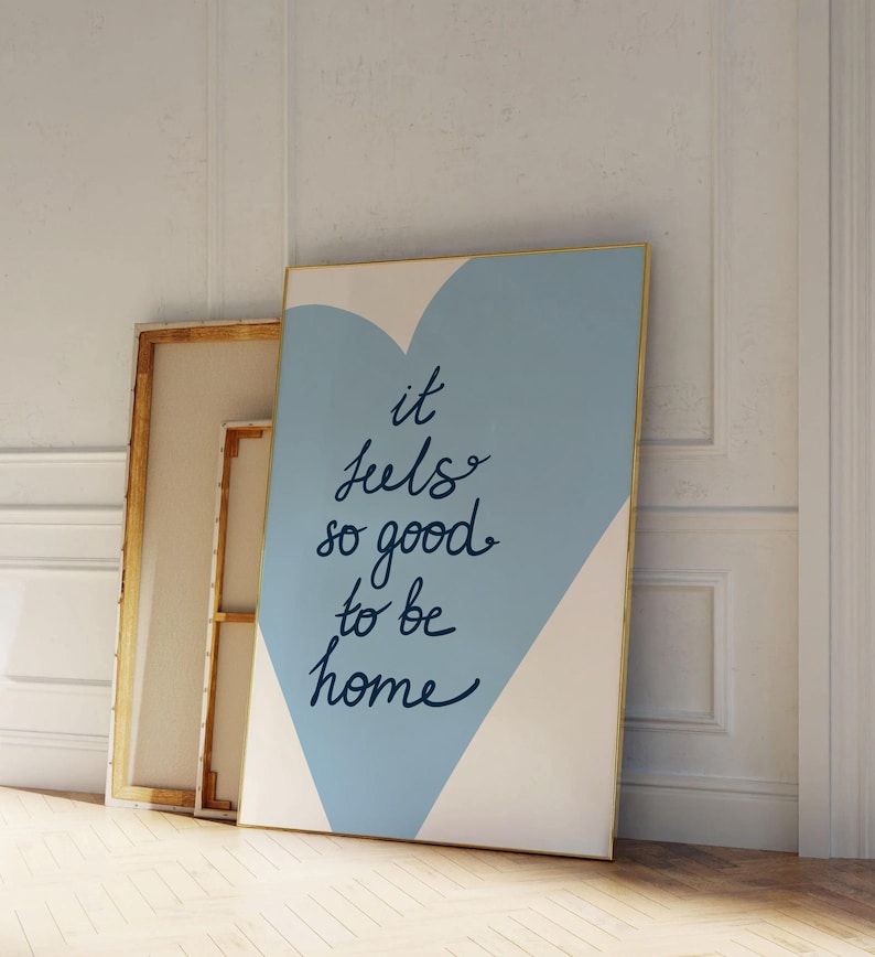 May include: A blue and white canvas print with a gold frame. The print features a blue heart shape with the text "it feels so good to be home" written in black.