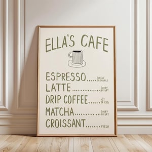 Puede incluir: Un cartel enmarcado con el texto "ELLA'S CAFE" en verde, con una ilustraci&oacute;n de una taza de caf&eacute;. El men&uacute; enumera opciones de espresso, latte, caf&eacute; de goteo, matcha y croissant. El cartel se muestra contra una pared blanca y un suelo de madera.