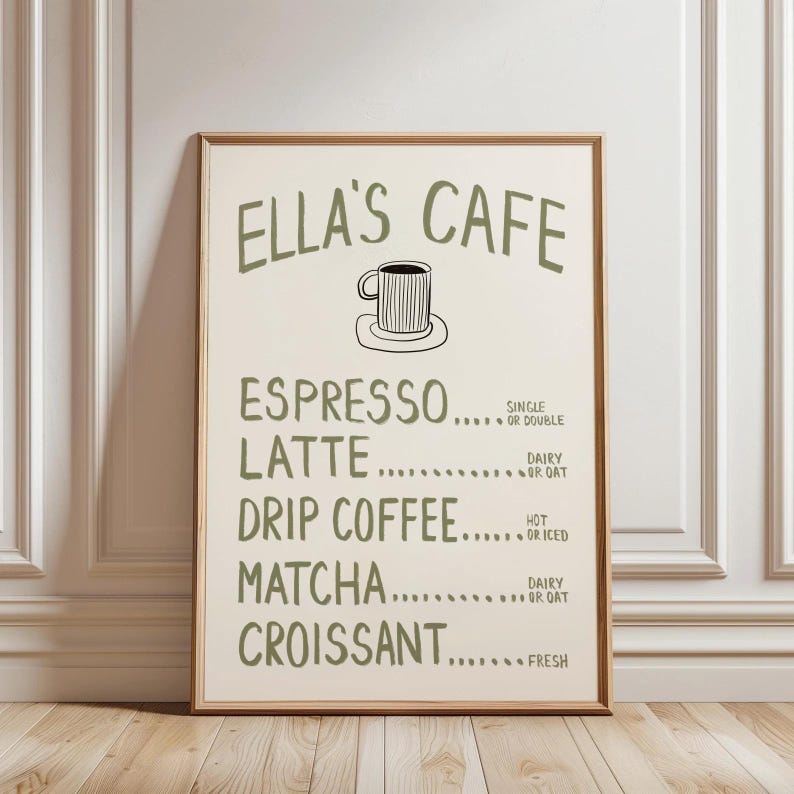 Puede incluir: Un cartel enmarcado con el texto "ELLA'S CAFE" en verde, con una ilustraci&oacute;n de una taza de caf&eacute;. El men&uacute; enumera opciones de espresso, latte, caf&eacute; de goteo, matcha y croissant. El cartel se muestra contra una pared blanca y un suelo de madera.