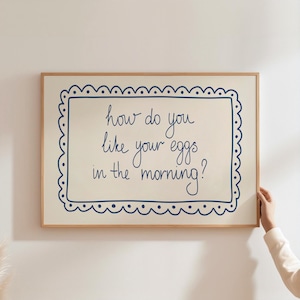 May include: A framed print with the text "how do you like your eggs in the morning?" in blue script. The text is inside a blue scalloped border on a cream background, set in a light wood frame.
