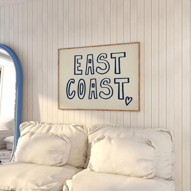 May include: A white framed print with the text "EAST COAST" in blue outlined letters with a small heart symbol. The print is hanging on a white wood panel wall above a white couch with two white pillows.