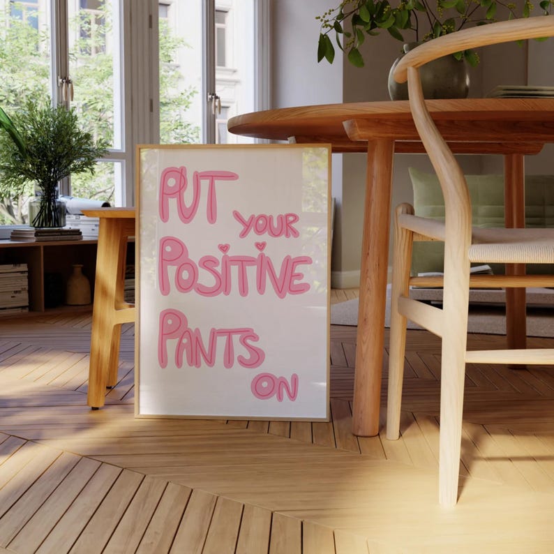 May include: A framed print with the text "PUT YOUR POSITIVE PANTS ON" in pink lettering on a white background. The print is hanging on a wall in a room with a wooden floor and a table and chair.