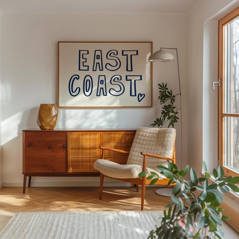 May include: A white framed poster with the text "EAST COAST" in blue lettering with a small heart symbol. The poster is hanging on a white wall above a wooden cabinet and a white armchair.