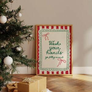 May include: A framed sign with the text "Wash your hands ya filthy animal" in green script. The sign has a red and white striped border and a green scalloped edge. A Christmas tree with ornaments is in the background.