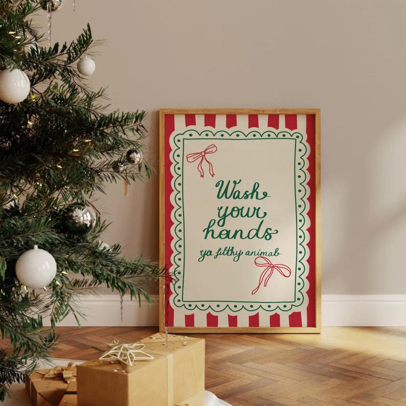 May include: A framed sign with the text "Wash your hands ya filthy animal" in green script. The sign has a red and white striped border and a green scalloped edge. A Christmas tree with ornaments is in the background.