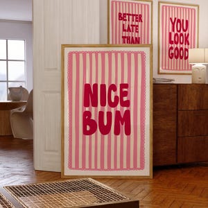 Op de afbeelding: Drie ingelijste kunstdrukken met roze en witte gestreepte achtergronden en tekst. De grootste print zegt "NICE BUM" in roze letters. De andere twee prints zeggen "BETTER LATE THAN" en "YOU LOOK GOOD". De frames zijn lichtbruin.