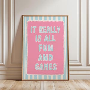 Puede incluir: Una impresión artística enmarcada con las palabras "IT REALLY IS ALL FUN AND GAMES" en azul claro sobre un fondo rosa. La impresión tiene un borde a rayas azul claro y blanco y está en un marco de madera.