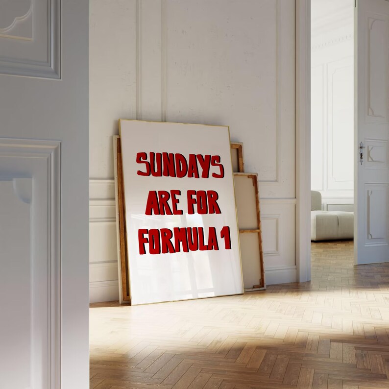 Op de afbeelding: Een wit canvas met de rode tekst "SUNDAYS ARE FOR FORMULA 1" in een lijst. Het kunstwerk staat tegen een witte muur in een kamer met een houten vloer. De kamer is goed verlicht.