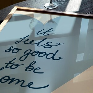 May include: A framed print with a blue and beige background. The print features the text "It feels so good to be home" in a dark blue cursive font.
