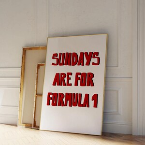 Op de afbeelding: Een wit ingelijste poster met de tekst "SUNDAYS ARE FOR FORMULA 1" in het rood, leunend tegen een muur. De poster heeft een gouden lijst. Achter de poster staat een ander ingelijst canvas.