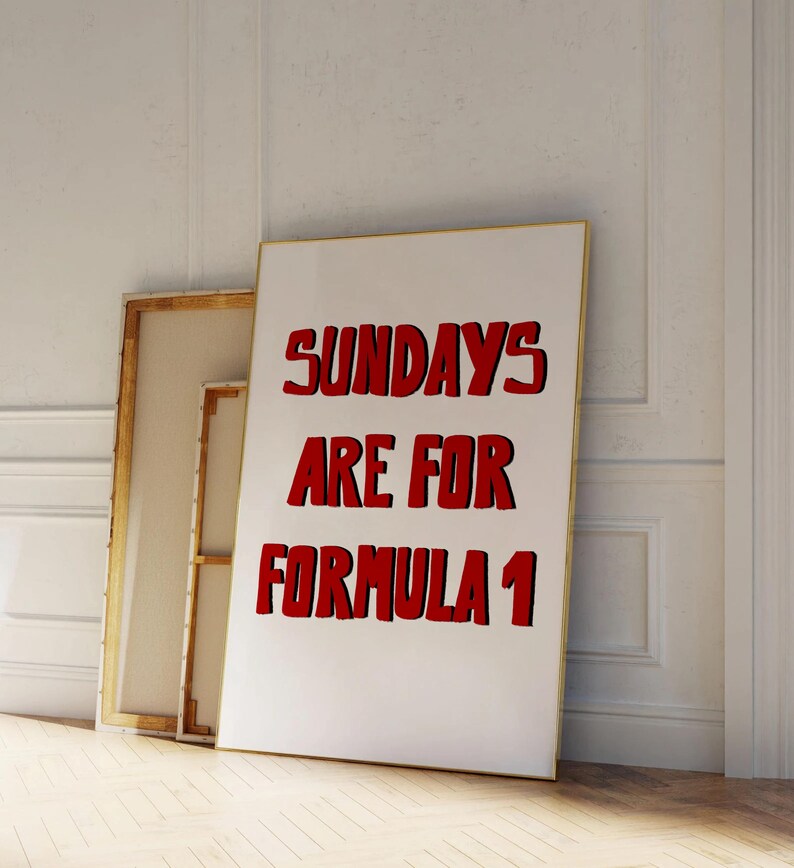 Op de afbeelding: Een wit ingelijste poster met de tekst "SUNDAYS ARE FOR FORMULA 1" in het rood, leunend tegen een muur. De poster heeft een gouden lijst. Achter de poster staat een ander ingelijst canvas.