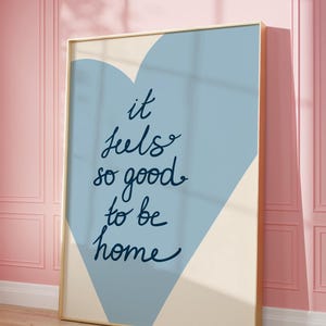 May include: A blue and cream colored poster with a heart shape in the background. The text on the poster reads "it feels so good to be home".