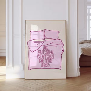 Puede incluir: Un cuadro enmarcado que representa una cama con almohadas y una manta en tonos rosas. El texto "NO OUTSIDE CLOTHES ON THE BED" está en la manta. La obra de arte está sobre un fondo neutro.