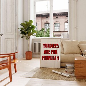 Op de afbeelding: Een ingelijste poster met de tekst "SUNDAYS ARE FOR FORMULA 1" in rode letters. De poster staat tegen een beige bank in een lichte kamer met een groot raam, een houten stoel en een potplant.