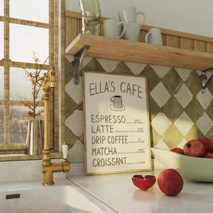 Puede incluir: Escena de cocina con un fregadero blanco, un grifo dorado y una estanter&iacute;a de madera con tazas y vasos blancos. Un letrero enmarcado dice "Ella's Cafe" con un men&uacute; de caf&eacute;. Granadas en la encimera. Una ventana es visible en el fondo.
