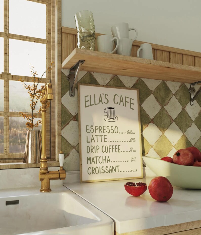 Puede incluir: Escena de cocina con un fregadero blanco, un grifo dorado y una estanter&iacute;a de madera con tazas y vasos blancos. Un letrero enmarcado dice "Ella's Cafe" con un men&uacute; de caf&eacute;. Granadas en la encimera. Una ventana es visible en el fondo.