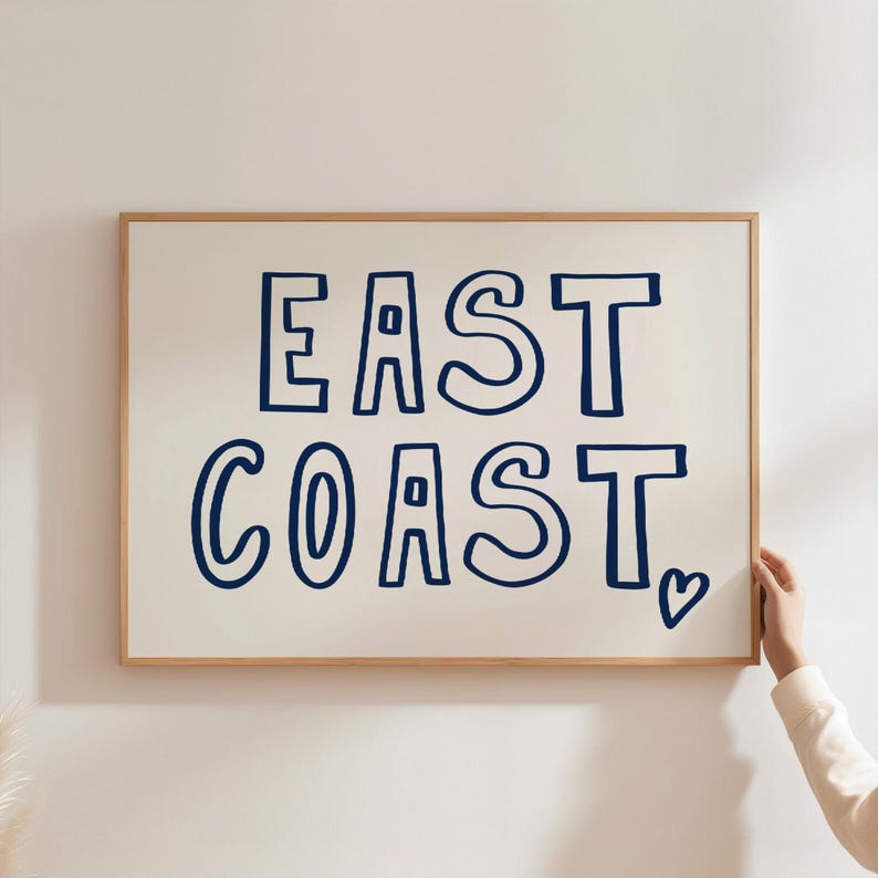 May include: A framed print with the text "EAST COAST" in a navy blue font with a heart symbol at the end. The print is on a white background and has a light wood frame.