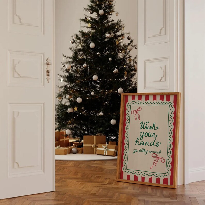 May include: A framed sign with the text "Wash your hands ya filthy animal" in green script, accented with red bows and a scalloped border. The sign is leaning against a wall near a decorated Christmas tree and wrapped gifts.