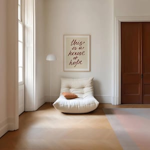 May include: A white, tufted, oversized chair with a brown throw pillow in front of a beige wall with a framed print that reads 'this is a house of hope' in red cursive lettering. The chair is in a room with white walls and a wooden floor.