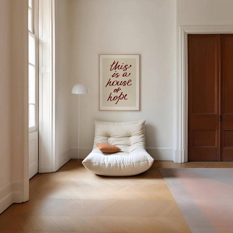 May include: A white, tufted, oversized chair with a brown throw pillow in front of a beige wall with a framed print that reads 'this is a house of hope' in red cursive lettering. The chair is in a room with white walls and a wooden floor.