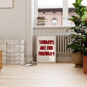 Op de afbeelding: Een ingelijste poster met de tekst "SUNDAYS ARE FOR FORMULA 1" in rode letters. De poster staat in een kamer met een raam, planten en een stapel tijdschriften. De kamer heeft een lichtgekleurde houten vloer.