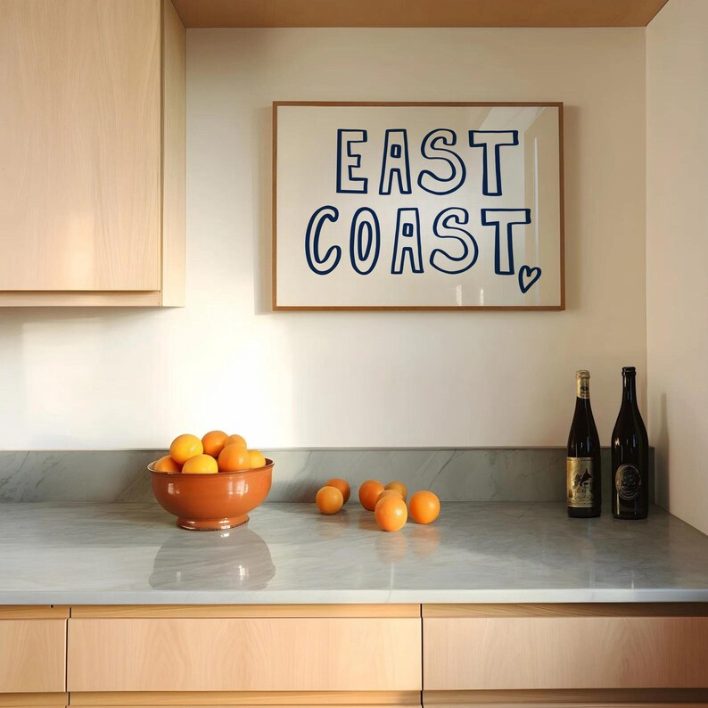May include: A kitchen counter with a bowl of oranges, two bottles of wine, and a framed print that says "EAST COAST" with a heart.