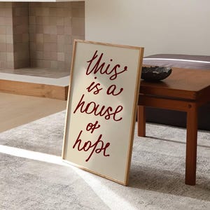 May include: A framed sign with the text 'This is a house of hope' written in red cursive on a cream background.