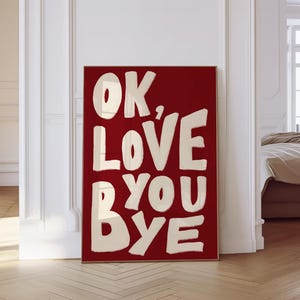Pode incluir: Uma impressão de arte emoldurada com fundo vermelho e as palavras "OK, LOVE YOU BYE" em letras grandes, brancas sujas, em estilo manuscrito. A obra de arte é exibida em uma sala com piso de parquet e paredes brancas.