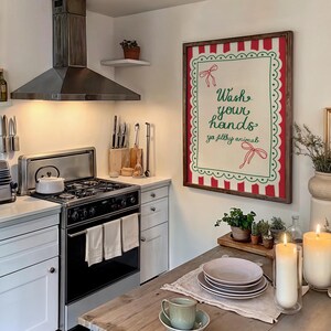 May include: A kitchen scene featuring a framed sign with the text "Wash your hands ya filthy animal." The sign has a red and green border. A stainless steel stove, range hood, and white cabinets are also visible. A wooden table is set with plates and a teacup.