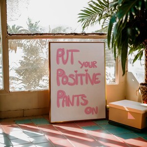 May include: A white framed poster with pink lettering that reads "Put your positive pants on".