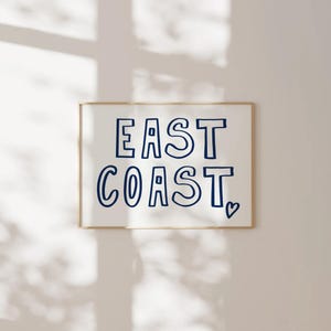 May include: A white framed print with the text "EAST COAST" in blue outlined letters with a small heart symbol at the bottom right corner.