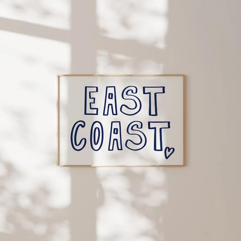 May include: A white framed print with the text "EAST COAST" in blue outlined letters with a small heart symbol at the bottom right corner.