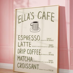Puede incluir: Un letrero enmarcado para Ella's Cafe, con un men&uacute; que incluye opciones como espresso, latte, caf&eacute; de goteo, matcha y croissant. El letrero es de color beige claro con texto verde oscuro y una ilustraci&oacute;n de taza de caf&eacute;. Est&aacute; apoyado contra una pared rosa.