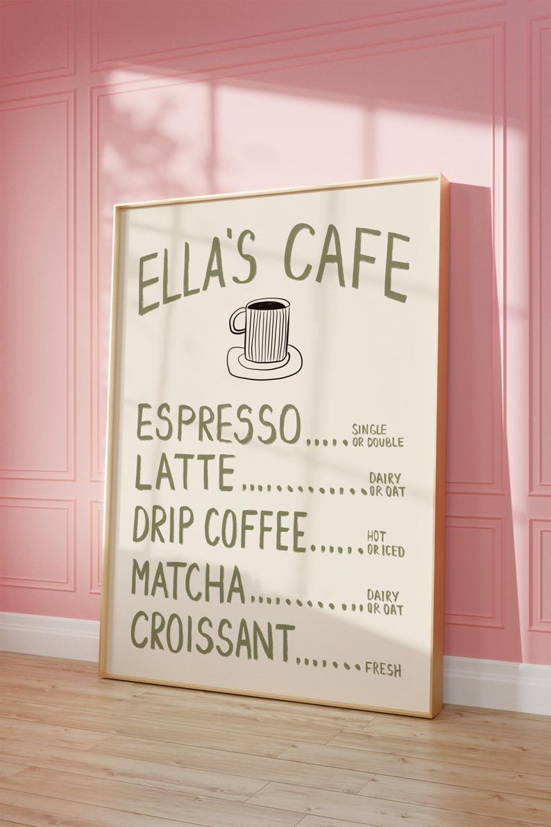 Puede incluir: Un letrero enmarcado para Ella's Cafe, con un men&uacute; que incluye opciones como espresso, latte, caf&eacute; de goteo, matcha y croissant. El letrero es de color beige claro con texto verde oscuro y una ilustraci&oacute;n de taza de caf&eacute;. Est&aacute; apoyado contra una pared rosa.