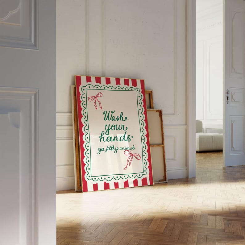 May include: A framed sign with a red and white striped border and the text "Wash your hands ya filthy animal" in green script. The sign has a scalloped green border and red bow accents. The sign is leaning against a wall in a room with a wooden floor.