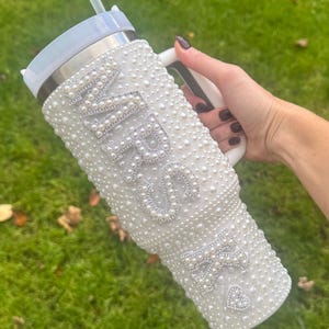 May include: White tumbler covered in faux pearls, with the word "MRS" and a heart-shaped design. The tumbler has a white handle and a straw. The background is green grass.