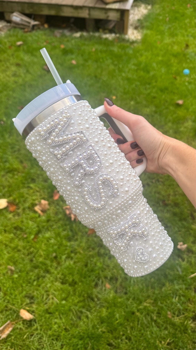 May include: White tumbler covered in faux pearls, with the word "MRS" and a heart-shaped design. The tumbler has a white handle and a straw. The background is green grass.