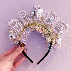 May include: A festive headband with the word "BRIDE" spelled out in white pearls and rhinestones. The headband features disco ball accents and iridescent tinsel, perfect for a bachelorette party or bridal celebration.